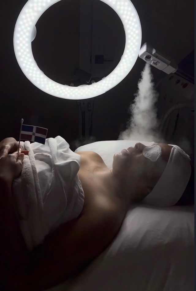 Client relaxing under a facial steamer and ring light during a facial at BellaFace